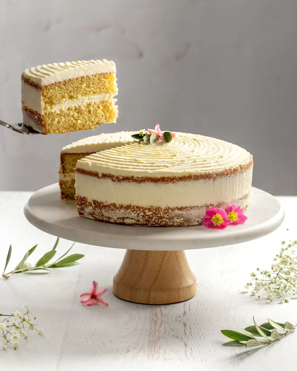 Naked vanilla cake on a white cake stand with a slice being removed, surrounded by flowers and greenery.