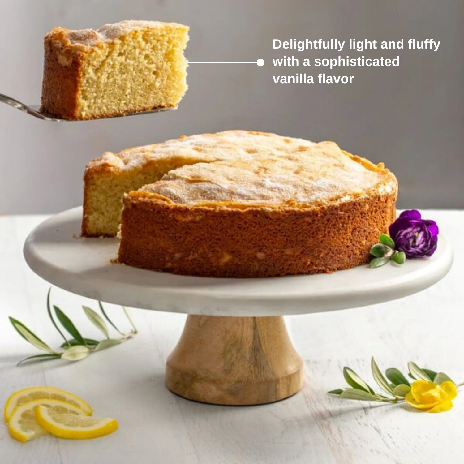Naked sugar crusted cake with a slice taken out on a white cake stand. Sliced lemons, flowers, and olive leaves surrounding on the table below.